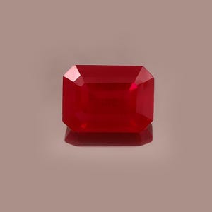 May include: A faceted, rectangular-cut ruby gemstone. The gem is a deep, saturated red color with a reflective surface. The gemstone is set against a neutral background, highlighting its brilliance and clarity. The cut is an emerald cut.