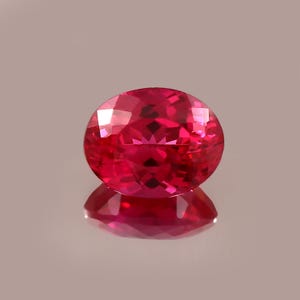 Puede incluir: Una gema de rubí tallada en forma ovalada y facetada. La gema es de un color rojo vibrante y saturado, con múltiples facetas que capturan la luz. El rubí se encuentra sobre un fondo neutro, con su reflejo visible debajo.