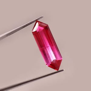 Puede incluir: Una gema rosa vibrante, facetada y alargada, es sostenida por unas pinzas plateadas. La gema tiene forma rectangular con extremos angulados, reflejando la luz. El fondo neutro resalta la claridad y el color de la gema.