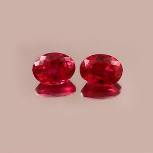 May include: Two oval-cut, faceted, bright red gemstones. The gems are displayed on a reflective surface, creating a mirrored effect. The stones have a vibrant, saturated color and a high degree of clarity, suggesting they are of good quality.