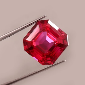 Può includere: Un rubino sfaccettato a taglio quadrato, tenuto da una pinzetta argentata. Il rubino è di un rosso intenso, con molteplici sfaccettature che riflettono la luce. La pinzetta tiene la gemma su uno sfondo neutro.