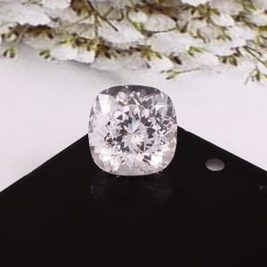 May include: A cushion-cut gemstone with a pale pink colour. The gem is set against a black surface, with a blurred floral arrangement in the background. The facets of the stone reflect light, creating a sparkling effect. Suitable for jewellery.