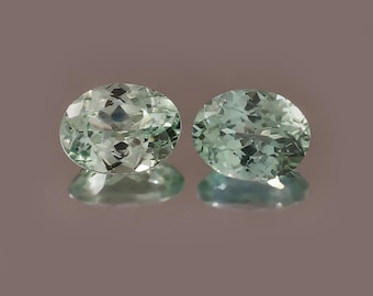 May include: Two oval-cut gemstones with a pale mint green hue and multifaceted surfaces. The stones are positioned side-by-side, displaying clear internal reflections and a brilliant, transparent quality that catches the light.