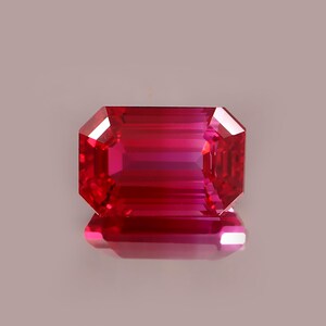 Op de afbeelding: Een levendige, rechthoekig geslepen robijn edelsteen. De edelsteen is diep rood met een reflecterend oppervlak, met interne facetten. De edelsteen is geplaatst tegen een neutrale achtergrond.