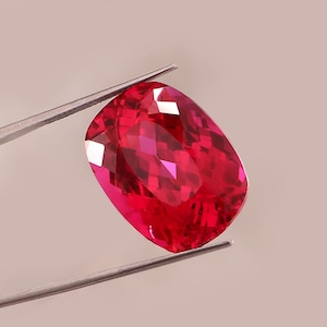 May include: A faceted, cushion-cut, red gemstone held by tweezers. The gem has a vibrant, deep red color with multiple facets that reflect light. The shape is rectangular with rounded corners, and the tweezers are silver.