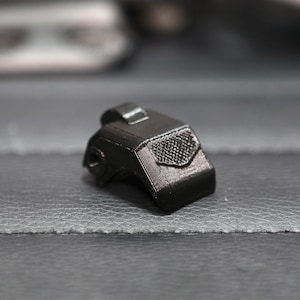 May include: A small, black, 3D-printed object with a textured surface and a rectangular shape. It features a small, raised section on top and a patterned design on the front. The object is resting on a dark gray surface.
