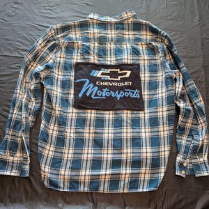 May include: Blue and white plaid button-down shirt with a black patch featuring the Chevrolet logo and the words "Motorsports" in blue. The shirt has long sleeves and a classic collar.