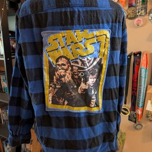 May include: A long-sleeve button-down shirt featuring a blue and black horizontal striped pattern. A rectangular patch is sewn onto the back, displaying the text STAR WARS and an illustration of Chewbacca and Han Solo with a gold glitter border.