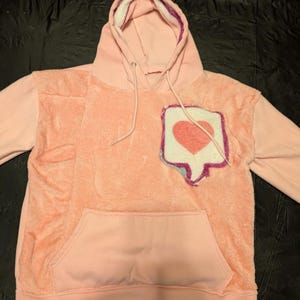 May include: A light pink hooded sweatshirt featuring a fuzzy, textured front panel and a large, stitched patch on the chest. The patch is shaped like a social media notification bubble with a pink heart icon, outlined in purple and blue thread.