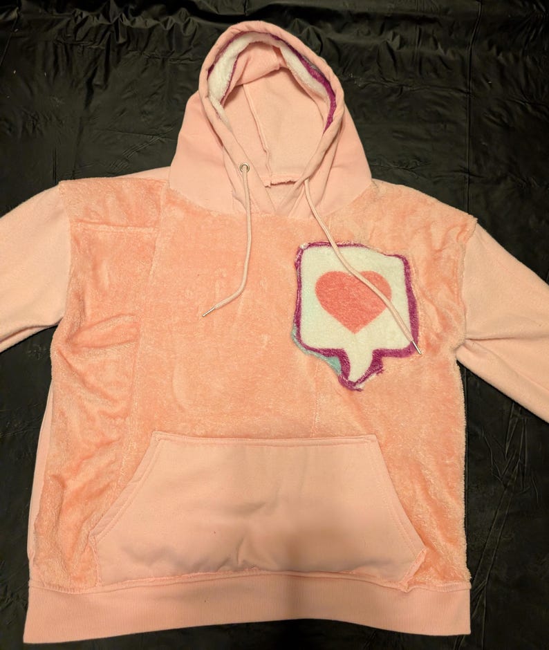 May include: A light pink hooded sweatshirt featuring a fuzzy, textured front panel and a large, stitched patch on the chest. The patch is shaped like a social media notification bubble with a pink heart icon, outlined in purple and blue thread.
