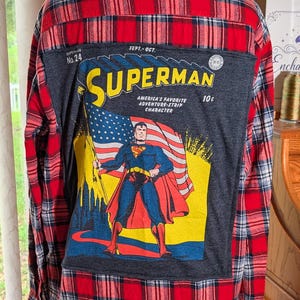 May include: A red, black, and white plaid long-sleeve button-down shirt featuring a large rectangular graphic on the back. The graphic depicts a vintage Superman comic book cover with the text Superman, No. 24, Sept.-Oct., and 10 cents.