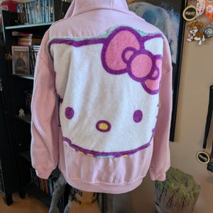 May include: A light pink hooded sweatshirt featuring a large, plush, white Hello Kitty character applique on the back. The character design includes a bright pink bow, purple outlines, two solid purple eyes, and a yellow oval nose on the fabric.