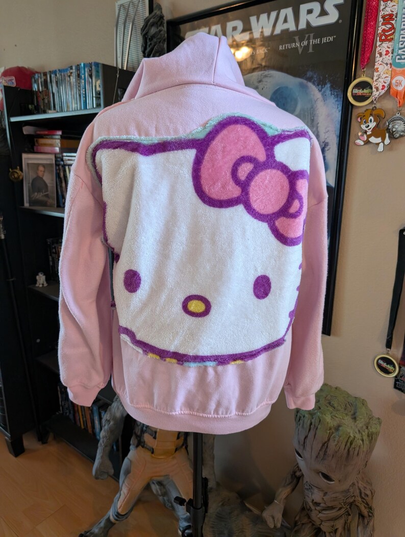 May include: A light pink hooded sweatshirt featuring a large, plush, white Hello Kitty character applique on the back. The character design includes a bright pink bow, purple outlines, two solid purple eyes, and a yellow oval nose on the fabric.