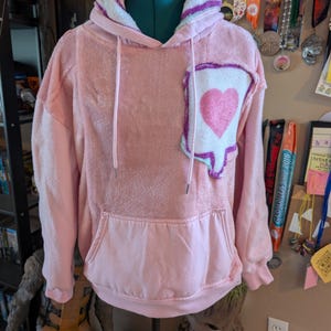 May include: A light pink fuzzy long-sleeve hoodie featuring a large white speech bubble patch on the left chest with a pink heart graphic inside. The hoodie includes a front kangaroo pocket, a drawstring hood with white lining, and ribbed cuffs.