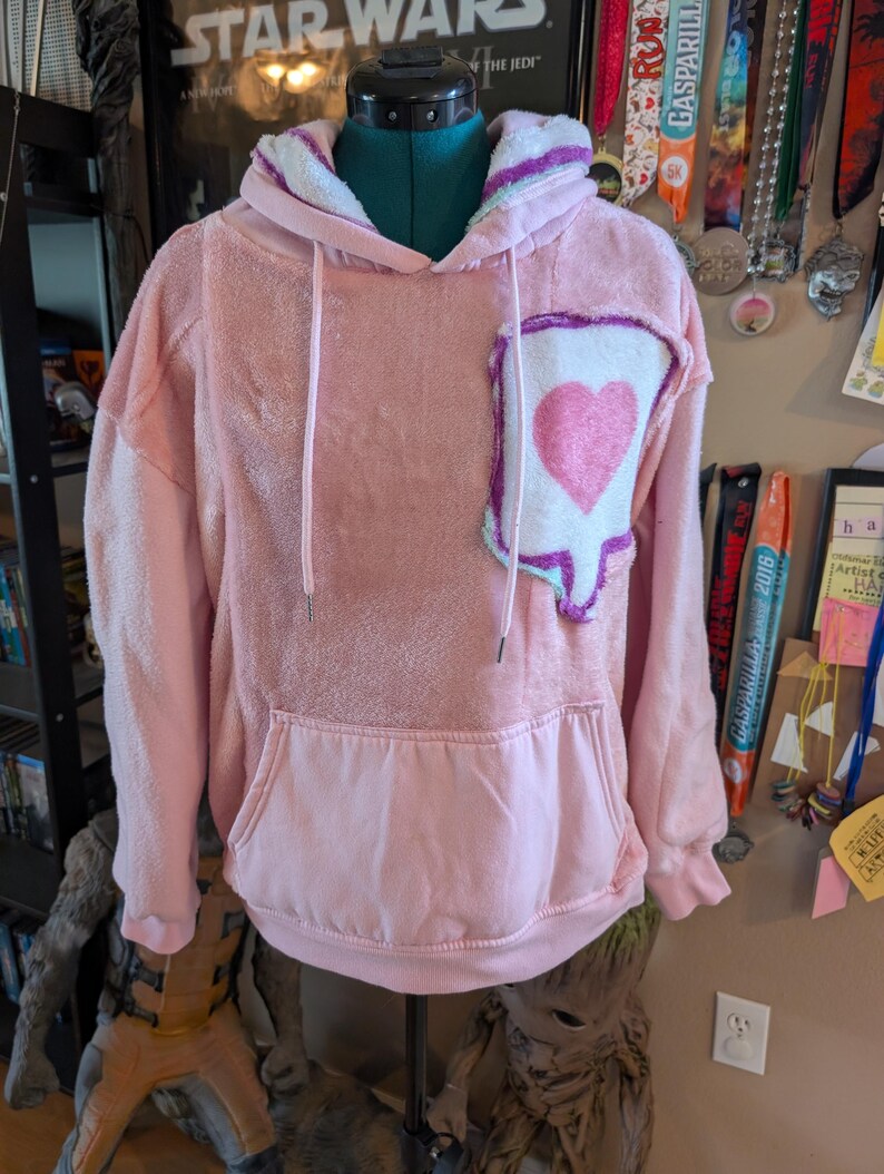 May include: A light pink fuzzy long-sleeve hoodie featuring a large white speech bubble patch on the left chest with a pink heart graphic inside. The hoodie includes a front kangaroo pocket, a drawstring hood with white lining, and ribbed cuffs.