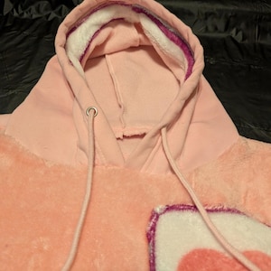 May include: A light pink plush hoodie featuring a hood with a white and purple inner lining, matching pink drawstrings with silver metal eyelets, and a soft, fuzzy texture throughout the garment with a partial heart graphic visible on the front.