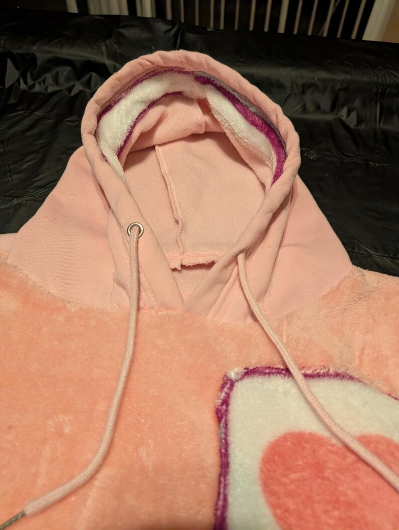May include: A light pink plush hoodie featuring a hood with a white and purple inner lining, matching pink drawstrings with silver metal eyelets, and a soft, fuzzy texture throughout the garment with a partial heart graphic visible on the front.