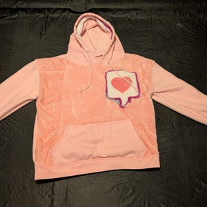 May include: A light pink long-sleeve hoodie featuring a textured, fuzzy central panel and a front kangaroo pocket. On the upper left chest, there is a patch shaped like a speech bubble containing a red heart graphic with a purple and blue border.