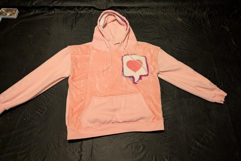 May include: A light pink long-sleeve hoodie featuring a textured, fuzzy central panel and a front kangaroo pocket. On the upper left chest, there is a patch shaped like a speech bubble containing a red heart graphic with a purple and blue border.