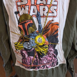 May include: An olive green jacket with a white patch featuring a Star Wars graphic. The graphic includes the Star Wars logo, Boba Fett, and other characters in shades of orange, yellow, pink, and blue.