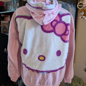 May include: A light pink hooded sweatshirt featuring a large, plush white applique of the Hello Kitty character on the back. The character design includes a pink bow, purple outlines, and yellow accents. The hoodie has long sleeves and a ribbed hem.