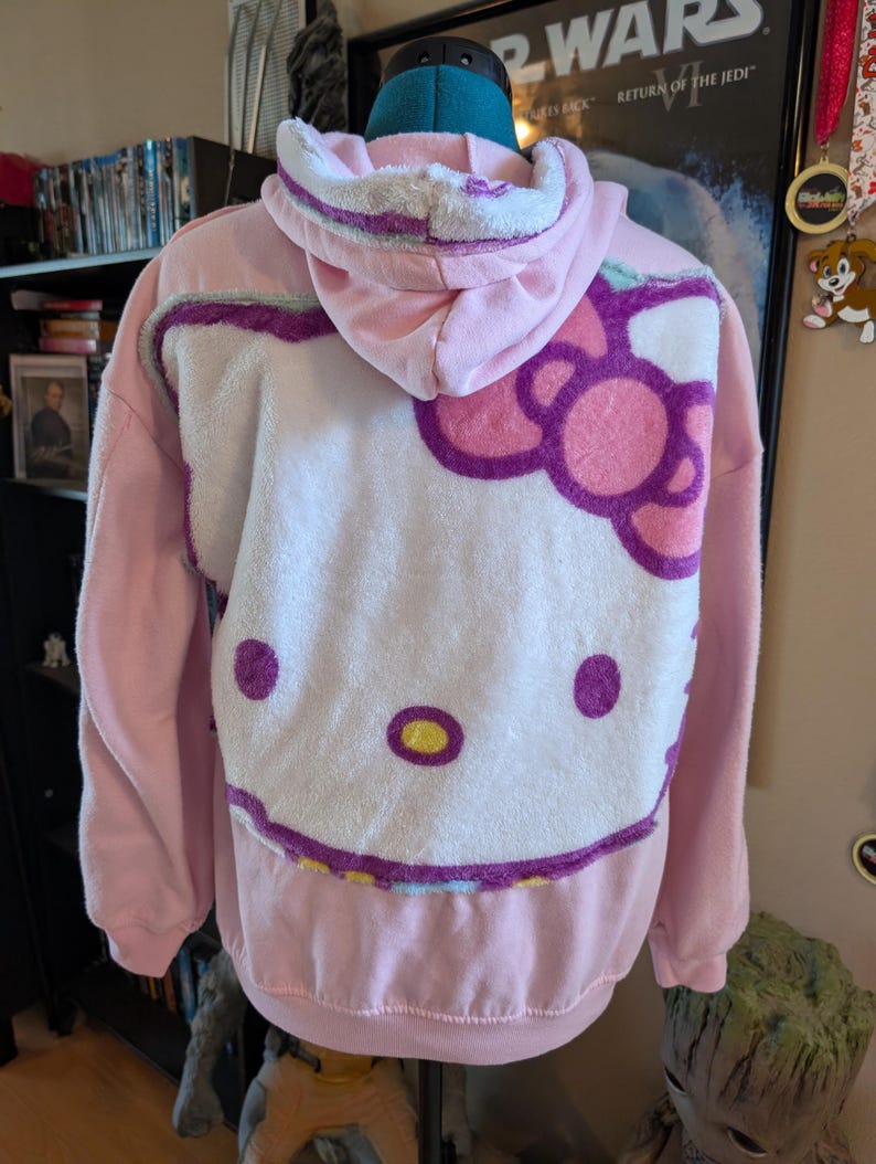 May include: A light pink hooded sweatshirt featuring a large, plush white applique of the Hello Kitty character on the back. The character design includes a pink bow, purple outlines, and yellow accents. The hoodie has long sleeves and a ribbed hem.