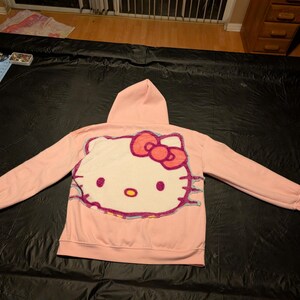 May include: A light pink hooded sweatshirt featuring a large, textured, white Hello Kitty character graphic on the back. The character has a bright red bow, black eyes, a yellow nose, and is outlined with a multi-colored, fuzzy, yarn-like border.