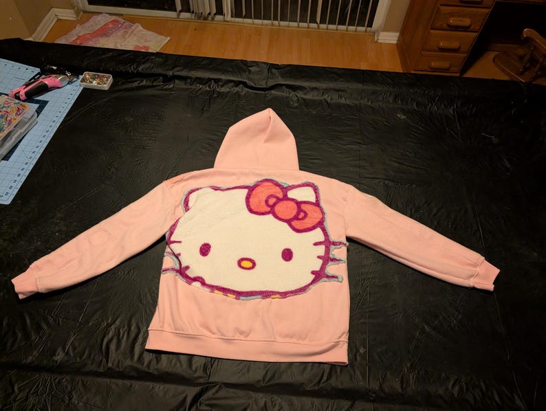 May include: A light pink hooded sweatshirt featuring a large, textured, white Hello Kitty character graphic on the back. The character has a bright red bow, black eyes, a yellow nose, and is outlined with a multi-colored, fuzzy, yarn-like border.