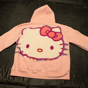 May include: A light pink hooded sweatshirt featuring a large, textured, tufted-style applique of the Hello Kitty character on the back. The character design includes a white face, a bright pink bow, black whiskers, and yellow nose details.