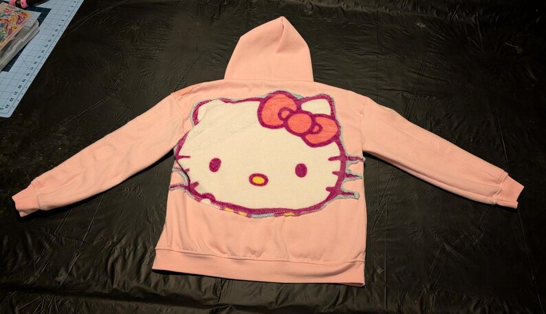 May include: A light pink hooded sweatshirt featuring a large, textured, tufted-style applique of the Hello Kitty character on the back. The character design includes a white face, a bright pink bow, black whiskers, and yellow nose details.