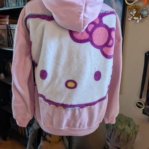 May include: A light pink hooded sweatshirt featuring a large, plush, white Hello Kitty character face appliqu&eacute; on the back. The character design includes a pink bow, purple outlines, and yellow accents on the nose, all rendered in a soft, textured fabric.