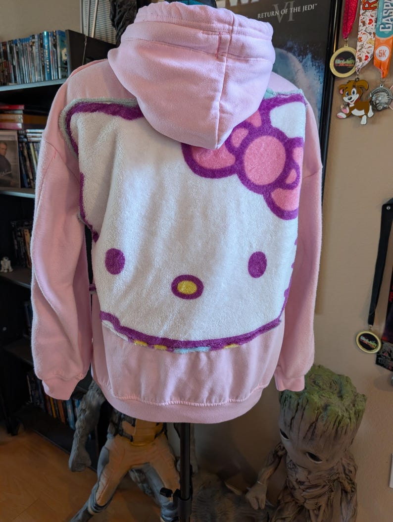 May include: A light pink hooded sweatshirt featuring a large, plush, white Hello Kitty character face appliqu&eacute; on the back. The character design includes a pink bow, purple outlines, and yellow accents on the nose, all rendered in a soft, textured fabric.
