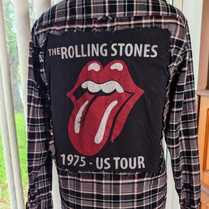 May include: A long-sleeve button-down shirt in a black, white, and red plaid pattern. The back features a large, rectangular black fabric patch with the text THE ROLLING STONES, a red tongue and lips graphic, and the text 1975 - US TOUR.