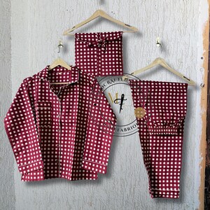 May include: A burgundy and white checkered pajama set, including a button-down shirt and pants, hangs on a wooden hanger. The set is displayed with a matching pair of pajama pants. The pajamas have white trim.