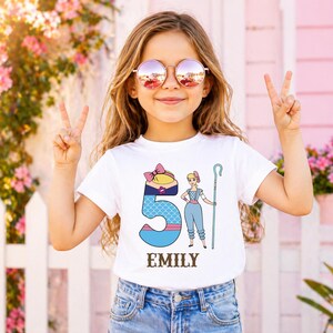 以下が含まれることがあります： 白いTシャツを着た若い女の子。Tシャツには、数字の5、漫画のキャラクター、そして「EMILY」という名前が描かれたカラフルなグラフィックがプリントされています。サングラスをかけ、両手でピースサインをしています。背景には白い柵とピンクの花があります。