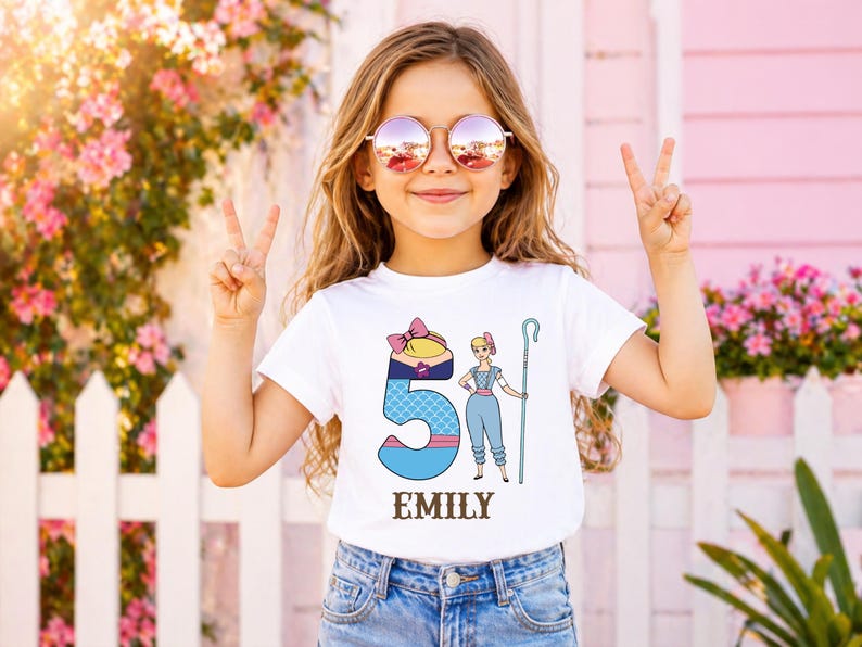 以下が含まれることがあります： 白いTシャツを着た若い女の子。Tシャツには、数字の5、漫画のキャラクター、そして「EMILY」という名前が描かれたカラフルなグラフィックがプリントされています。サングラスをかけ、両手でピースサインをしています。背景には白い柵とピンクの花があります。