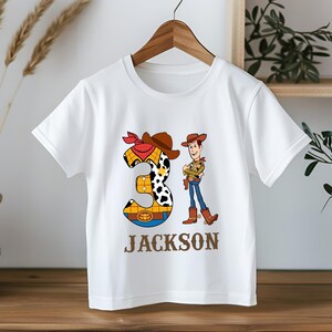 以下が含まれることがあります： 白いTシャツ。トイ・ストーリーのウッディの漫画のグラフィックと数字の3が特徴です。数字の3はカウボーイをテーマにしたデザインで、名前の「JACKSON」は茶色の文字で下に印刷されています。木製のハンガーにかかっています。