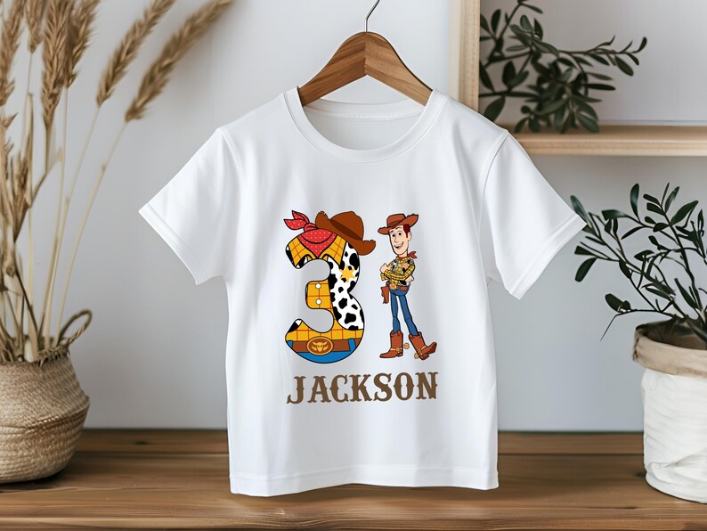 以下が含まれることがあります： 白いTシャツ。トイ・ストーリーのウッディの漫画のグラフィックと数字の3が特徴です。数字の3はカウボーイをテーマにしたデザインで、名前の「JACKSON」は茶色の文字で下に印刷されています。木製のハンガーにかかっています。
