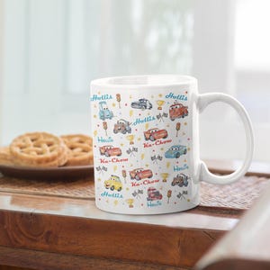 Puede incluir: Taza de cer&aacute;mica blanca decorada con coches de dibujos animados coloridos, trofeos y rayos. La taza presenta los textos "Hollis" y "Ka-Chow" en azul y rojo. La taza est&aacute; sobre una mesa de madera con galletas.