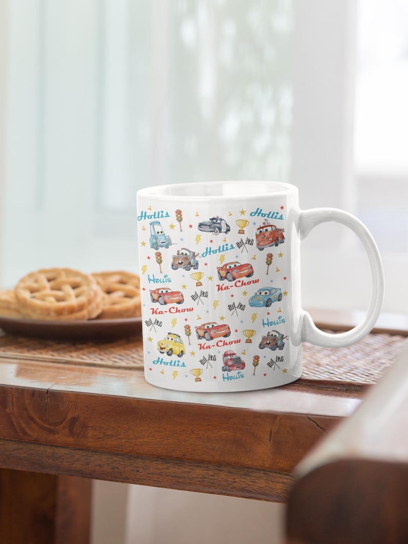 Puede incluir: Taza de cer&aacute;mica blanca decorada con coches de dibujos animados coloridos, trofeos y rayos. La taza presenta los textos "Hollis" y "Ka-Chow" en azul y rojo. La taza est&aacute; sobre una mesa de madera con galletas.