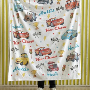 Puede incluir: Manta blanca con ilustraciones de coches de dibujos animados, trofeos, rayos y estrellas. La manta tiene el nombre "Hollis" y la frase "Ka-Chow" impresa. Los coches son de varios colores, incluyendo rojo, azul y amarillo.