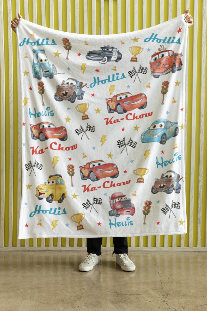 Puede incluir: Manta blanca con ilustraciones de coches de dibujos animados, trofeos, rayos y estrellas. La manta tiene el nombre "Hollis" y la frase "Ka-Chow" impresa. Los coches son de varios colores, incluyendo rojo, azul y amarillo.