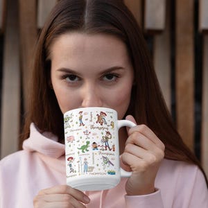 Puede incluir: Taza de cer&aacute;mica blanca con personajes de dibujos animados coloridos y el nombre "Thompson" en una fuente l&uacute;dica. La taza tiene un asa blanca y la sostiene una persona con una sudadera con capucha rosa. El fondo es una pared de madera.