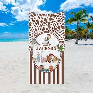 May include: A beach towel featuring a brown and white cowhide pattern at the top and brown vertical stripes at the bottom. The center has a circular design with cartoon characters and the name "Jackson". The towel is hanging on a clothesline on a sunny beach.