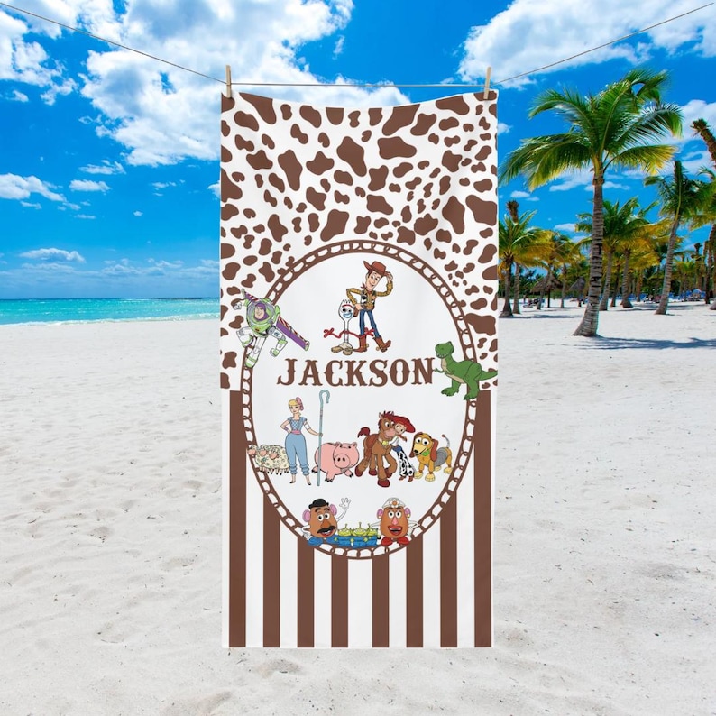 May include: A beach towel featuring a brown and white cowhide pattern at the top and brown vertical stripes at the bottom. The center has a circular design with cartoon characters and the name "Jackson". The towel is hanging on a clothesline on a sunny beach.