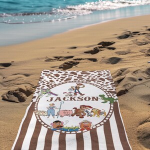 May include: A personalized beach towel with a brown and white cowhide pattern border and vertical brown stripes. The center features a circular design with cartoon characters and the name "JACKSON" in green letters.