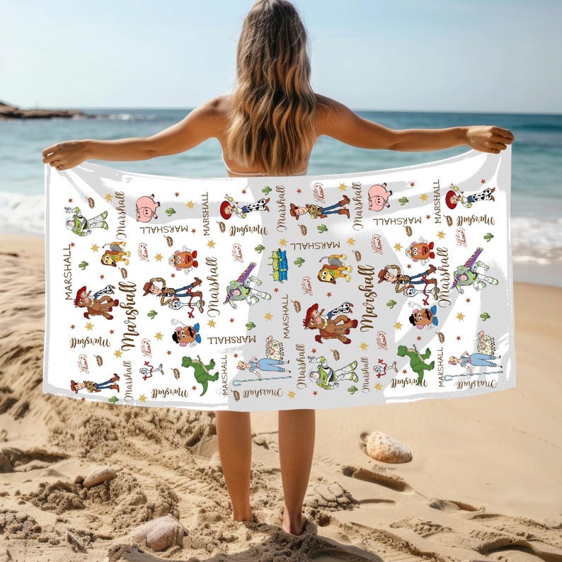 May include: White beach towel with a repeating pattern of cartoon characters and the name "Marshall". The towel is held up on a sandy beach with the sea in the background. The towel is rectangular in shape.