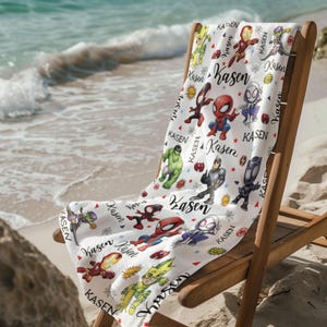 Pu&ograve; includere: Telo mare bianco con illustrazioni di supereroi dei cartoni animati e il nome "Kasen" in corsivo. L'asciugamano &egrave; appoggiato su una sedia a sdraio di legno su una spiaggia sabbiosa.