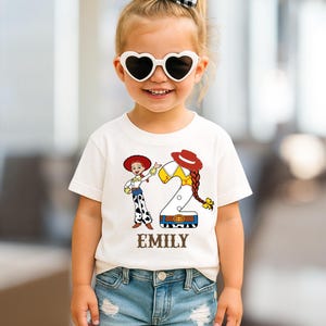 以下が含まれることがあります： 白いTシャツには、カウガールの漫画のグラフィックと数字の2が描かれています。「EMILY」という名前が下に印刷されています。子供はハート型のサングラスと青いジーンズを着用しています。