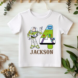 以下が含まれることがあります： 白いTシャツ。バズ・ライトイヤーの漫画の画像と、緑、青、白の数字4が描かれています。「JACKSON」という名前が茶色でプリントされています。木製のハンガーにTシャツが掛かっています。